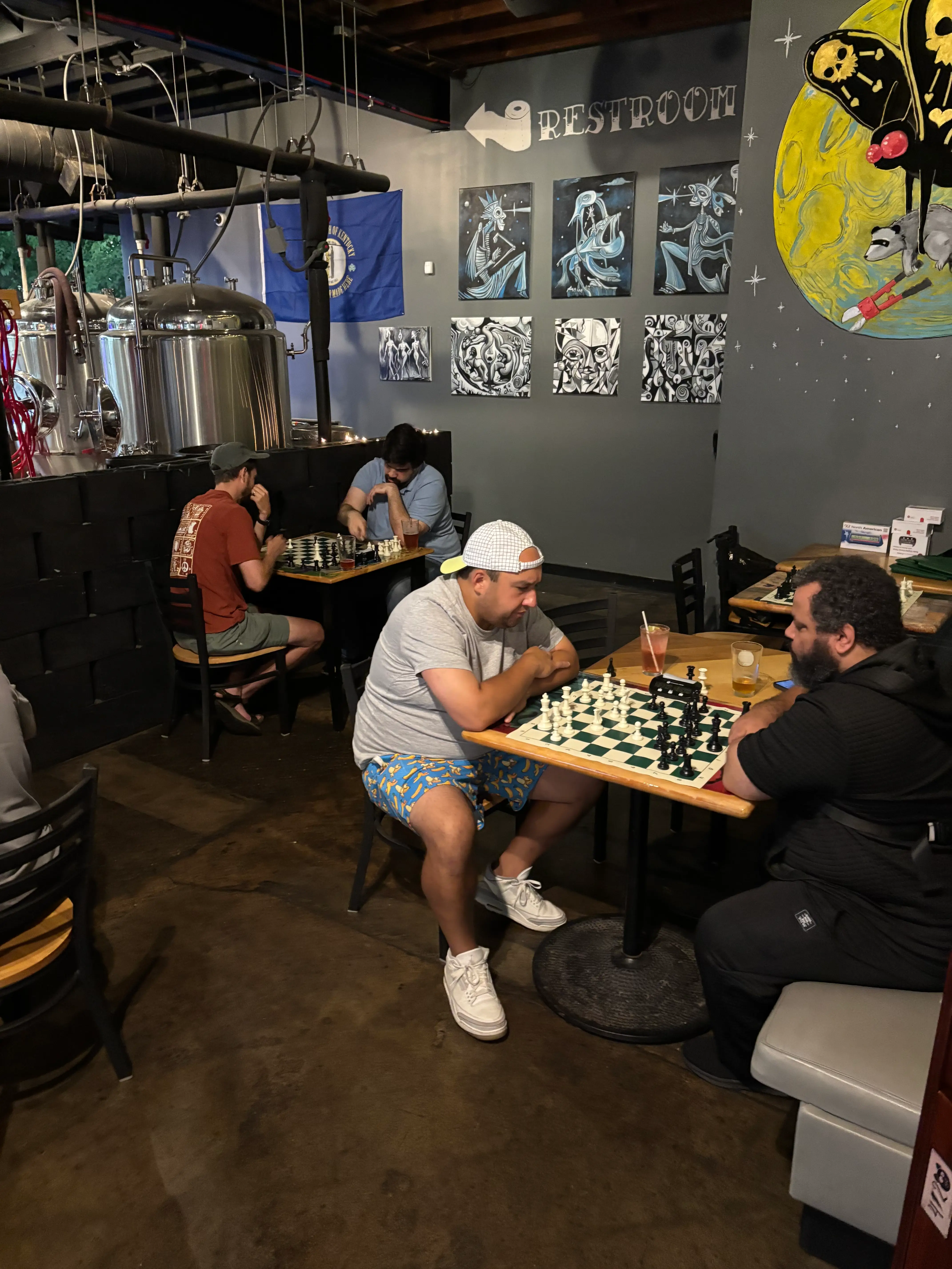 Four people playing chess at separate tables with brewery tanks and hanging art in the background.
