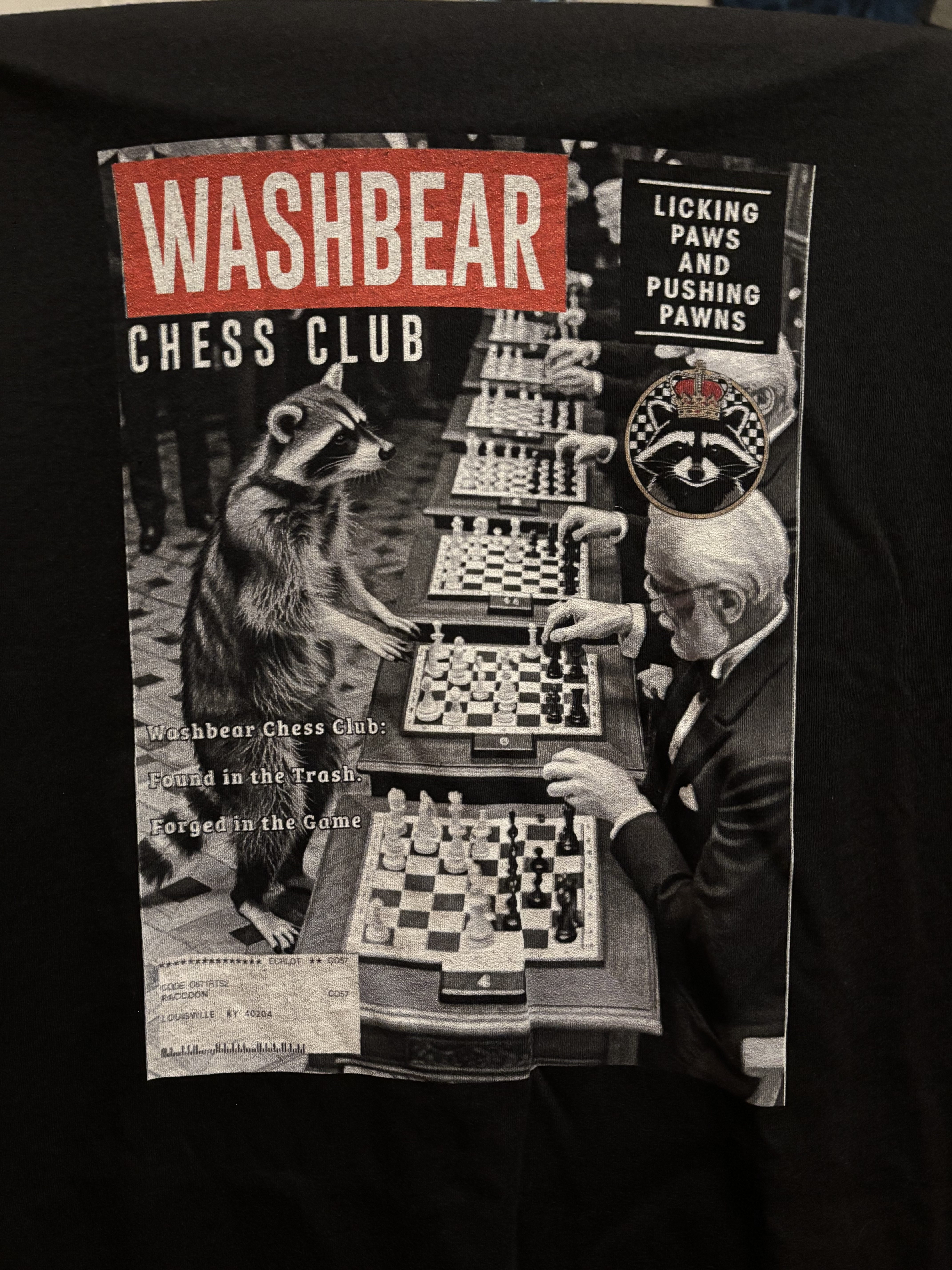 A black t-shirt with the design of a fake magazine featuring a raccoon playing a simul chess exhibition on the cover.